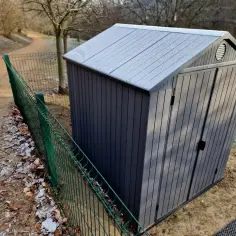 Szary domek narzędziowy na granicy działki, z zielonym ogrodzeniem. Budowa domu w granicy działki wymaga zgody sąsiada.