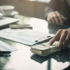 Dłoń sięga po stos banknotów leżący na stole obok laptopa i dokumentów.