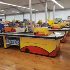 Kasy w Biedronce, gdzie pracownik zarabia na codzienne niskie ceny. W tle półki z produktami i żółte balony.
