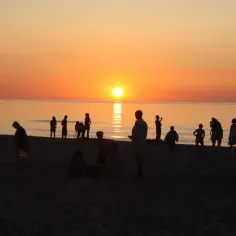 Zachód słońca w Sarbinowie. Ludzie spacerują po plaży, podziwiając pomarańczowe niebo i odbijające się w wodzie słońce.