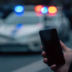 Dłoń trzyma smartfon, w tle migające światła policyjnego radiowozu. Czy to początek inwigilacji?