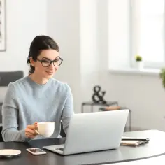 Kobieta z kubkiem kawy analizuje na laptopie, ile bierze biuro nieruchomości za sprzedaż działki.