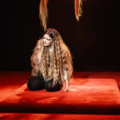 Aktorka na czerwonej scenie w teatrze w Warszawie, w błyszczącej kreacji i z długimi włosami, przeżywa dramatyczny moment.