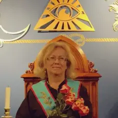 Kobieta w loży masońskiej, siedząca na ozdobnym tronie, z różami w dłoni. W tle symbole: oko opatrzności, słońce i księżyc.