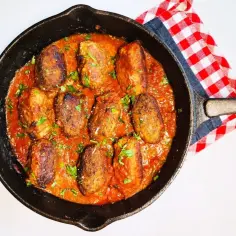 Hreczanyki w sosie pomidorowym, podane w żeliwnej patelni, ozdobione natką pietruszki. Obok czerwono-biała kratkowana serwetka.