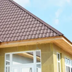 Nowy dach z brązowej blachodachówki, widoczne przedłużenie okapu dachu nad oknami i ścianą z izolacją.