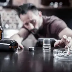 Mężczyzna w rozpaczy, otoczony butelkami i popielniczką z niedopałkiem. Nexpram i alkohol to jego towarzysze.
