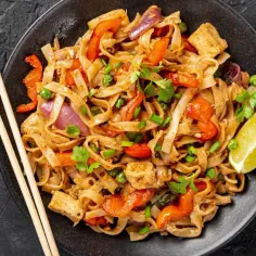 Talerz pełen makaronu ryżowego z warzywami, tofu i kolendrą, polany aromatycznym sosem pad thai. Obok pałeczki i ćwiartka limonki.