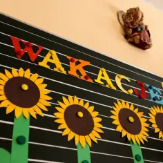 Kolorowe litery "WAKACJE!" i słoneczniki na tablicy zapowiadają radosne zakończenie roku szkolnego.