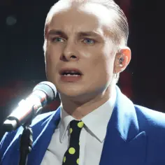 Ralph Kaminski w niebieskiej marynarce i krawacie w kropki śpiewa do mikrofonu.