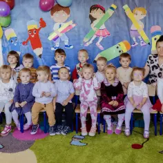 Grupa przedszkolaków z panią, w tym Tymon i Tymoteusz, pozuje do zdjęcia na tle kolorowych kredek.