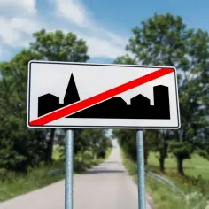 Znak D42: koniec obszaru zabudowany. Droga prowadzi przez zielone tereny, a na znaku sylwetka miasta przekreślona czerwoną linią.