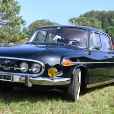 Lśniąca, czarna Tatra 603 z lat 60. na trawie, z chromowanymi detalami i okrągłymi reflektorami.