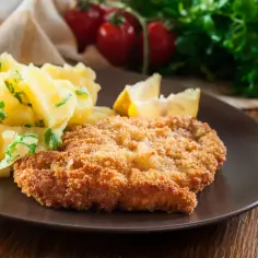 Kotlet schabowy w złocistej panierce z ziemniakami i cytryną na brązowym talerzu.