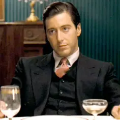 Młody Al Pacino w eleganckim garniturze, z papierosem w dłoni, siedzi przy stole z kieliszkami.