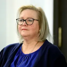 Kobieta w okularach, w granatowej bluzce i marynarce, z naszyjnikiem. Jej spojrzenie sugeruje skupienie, jakby czekała na kontrasygnatę.
