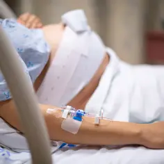 Kobieta w szpitalnym łóżku, z wenflonem w ręce, przechodzi trudny czas urlopu macierzyńskiego po poronieniu.