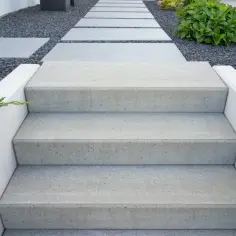 Betonowe schody zewnętrzne z jasnymi stopniami i białymi podstopnicami, otoczone żwirem i zielenią.