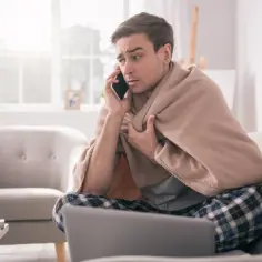 Chory mężczyzna owinięty kocem rozmawia przez telefon, szukając informacji, jak obliczyć świadczenie chorobowe.