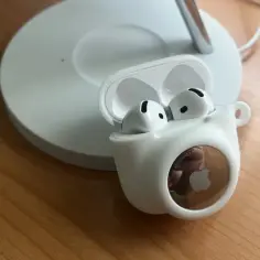 AirPods w białym etui z przyczepionym AirTagiem na drewnianym blacie.