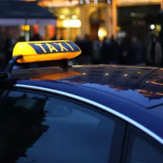 Żółty znak TAXI na dachu ciemnego samochodu, wieczorem, z rozmytym tłem miasta.