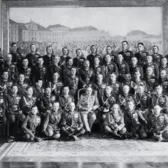 Grupa mężczyzn w mundurach wojskowych, być może z obozu zwolenników Piłsudskiego, pozuje do zdjęcia na tle obrazu.