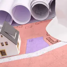 Model domu na planach, biały kask budowlany i zwoje dokumentacji. Rozważanie służebności gruntowej przy planowaniu inwestycji.