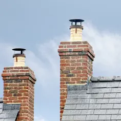 Dwa ceglane kominy z metalowymi nasadkami na tle błękitnego nieba z chmurami.