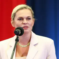 Minister edukacji przemawia do mikrofonu na tle czerwono-niebieskiej kurtyny.