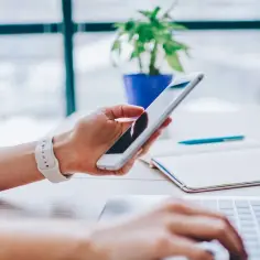 Dłoń trzyma smartfon, korzystając z tetheringu, podczas gdy druga ręka pisze na laptopie. Obok leży notatnik i długopis.