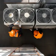 Dwóch techników w pomarańczowych kamizelkach i kaskach pracuje przy dużych wentylatorach klimatyzacji, być może sprawdzając, jak zmienić kierunek obrotu silnika jednofazowego.