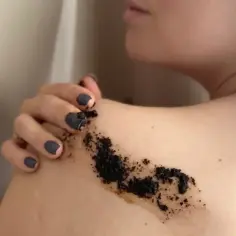 Kobieta aplikuje na ramię peeling z kawy, który ma ciemny, ziarnisty kolor.