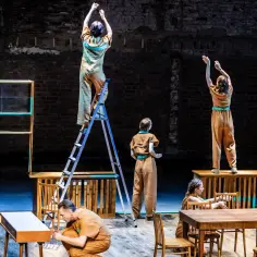 Scena teatralna: ludzie w brązowych strojach, jeden na drabinie, inni przy stołach. Czy to apoteoza?
