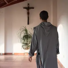 Mnich w szarym habicie idzie korytarzem pod krzyżem.