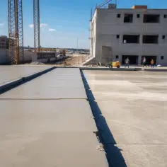 Świeżo wylany beton na ławy fundamentowe. W tle budowa, dźwigi i budynki.