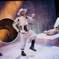 Aktor w białym stroju i kapeluszu tańczy na scenie. W tle, na podwyższeniu, klęczy druga postać. To scena z przedstawienia "Mały Książę teatr".