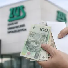 Ręce trzymają polskie banknoty przed budynkiem ZUS. Kiedy najlepiej przejść na emeryturę w 2026 roku?