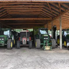 Drewniana wiata bez pozwolenia dla rolnika chroni cztery traktory, w tym zielone John Deere i czerwony Zetor, gotowe do pracy na polu.