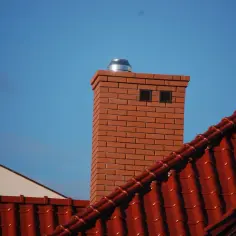 Ceglany komin z metalową nasadką na tle błękitnego nieba, obok czerwonego dachu.
