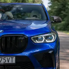 Błyszczący, niebieski BMW X5 M Competition na drodze leśnej. Charakterystyczne reflektory i czarny grill podkreślają jego sportowy charakter.