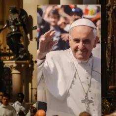 Papież Franciszek w białej szacie z krzyżem, na tle wnętrza kościoła.