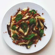 Makaron penne z suszonymi pomidorami, zielonymi liśćmi i pokruszonym tofu na białym talerzu.