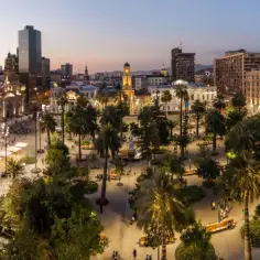 Wieczorny widok na plac w Santiago de Chile, z palmami, budynkami i ludźmi spacerującymi po oświetlonym deptaku.