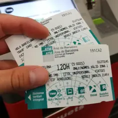 Dłoń trzyma bilety na komunikację miejską w Barcelonie. Na biletach widnieje napis "Hola Barcelona Travel Card" oraz informacje o ważności.