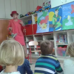 Nauczycielka w czerwonym kapeluszu i stroju opowiada bajkę, trzymając koszyczek Czerwonego Kapturka. Dzieci słuchają z uwagą.