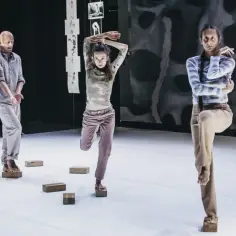 Trzech tancerzy wykonuje skomplikowaną choreografię na scenie, balansując na drewnianych klockach.