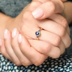 Złoty pierścionek z kamieniem, otoczony mniejszymi kamieniami, ozdabia dłoń z delikatnym różowym manicure.