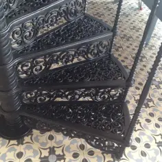 Stare, czarne schody kręcone z kutego żelaza z ozdobnymi, ażurowymi stopniami, stoją na wzorzystej, zabytkowej posadzce.