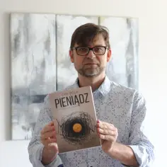 Mężczyzna w okularach trzyma powieść Emila Zoli "Pieniądz", której okładka przedstawia gniazdo z monetą.
