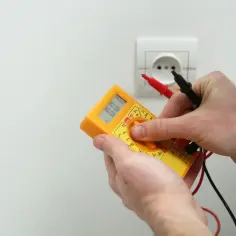 Ręce trzymające żółty multimetr z sondami przy białym gniazdku elektrycznym.